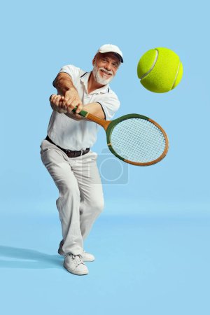 Téléchargez les photos : Returning ball. Portrait of handsome senior man in stylish white outfit playing tennis over blue background. Concept of leisure activity, hobby, lifestyle, fitness, emotions, retro style - en image libre de droit