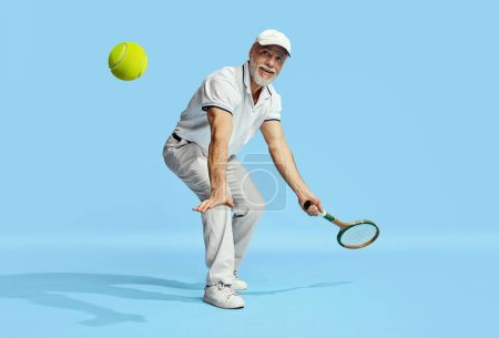 Téléchargez les photos : Happy look. Portrait of handsome senior man in stylish white outfit playing tennis over blue background. Concept of leisure activity, hobby, lifestyle, fitness, emotions, retro style - en image libre de droit