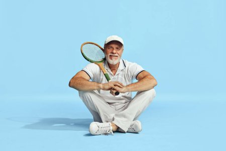 Téléchargez les photos : Delightful. Portrait of handsome senior man in stylish white outfit sitting, posing with tennis racket on blue background. Concept of leisure activity, hobby, lifestyle, fitness, emotions, retro style - en image libre de droit