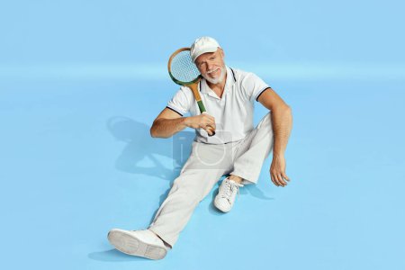 Téléchargez les photos : Relaxation. Portrait of handsome senior man in stylish white outfit sitting, posing with tennis racket on blue background. Concept of leisure activity, hobby, lifestyle, fitness, emotions, retro style - en image libre de droit
