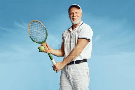 Téléchargez les photos : Portrait of handsome senior man in stylish white outfit posing with tennis racket over blue sky background. Concept of leisure activity, hobby, lifestyle, fitness, emotions, retro style - en image libre de droit