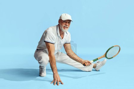 Téléchargez les photos : Portrait of handsome senior man in stylish white outfit sitting, posing with tennis racket over blue background. Concept of leisure activity, hobby, lifestyle, fitness, emotions, retro style - en image libre de droit