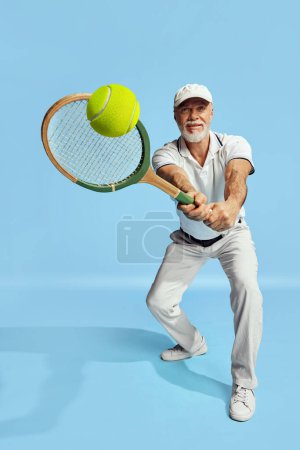 Téléchargez les photos : Serving ball. Portrait of handsome senior man in stylish white outfit playing tennis over blue background. Concept of leisure activity, hobby, lifestyle, fitness, emotions, retro style - en image libre de droit