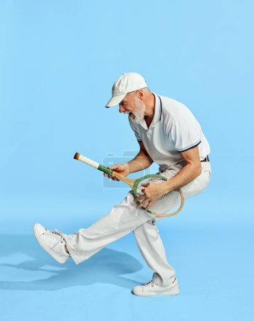 Téléchargez les photos : Portrait of handsome senior man in stylish white outfit posing with tennis racket on blue background. Playing like guitar. Concept of leisure activity, hobby, lifestyle, fitness, emotions, retro style - en image libre de droit
