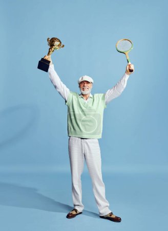 Téléchargez les photos : Winner. Portrait of handsome senior man in stylish outfit posing with tennis racket and trophy over blue background. Concept of leisure activity, hobby, lifestyle, fitness, emotions, retro style - en image libre de droit