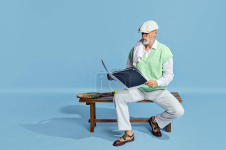 Téléchargez les photos : Portrait of handsome, serious, senior man in stylish outfit sitting on bench with sportive journal over blue background. Concept of leisure activity, hobby, lifestyle, fitness, emotions, retro style - en image libre de droit