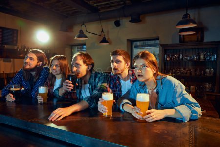 Foto de Group of young people, men and women, sport fans watching match at pub. Attentively looking, emotionally cheering up favourite team. Competition, championship, match translation. Emotions. Leisure - Imagen libre de derechos