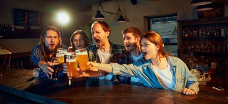 Foto de Group of young people, men and women, Fans emotionally cheering up favourite sport team at the pub. Clinking beer glasses. Successful game. Competition, championship, match translation. Emotions. - Imagen libre de derechos