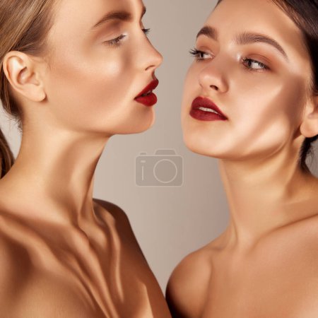 Téléchargez les photos : Well-kept skin. Portrait of two young, beautiful girls with red lips makeup isolated over grey studio background. Shadows. Concept of skincare, cosmetology, natural beauty, youth, spa, cosmetics. Ad - en image libre de droit