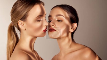 Foto de Clear, fresh, young face, skin. Portrait of two young, beautiful girls with red lips isolated over grey background. Shadows. Concept of skincare, cosmetology, natural beauty, youth, spa, cosmetics. Ad - Imagen libre de derechos