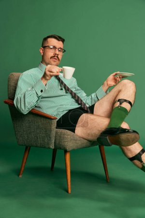 Foto de Morning coffee. Portrait of handsome man, businessman in shirt and underwear sitting on chair with serious face over green background. Concept of emotions, business, occupation, facial expression - Imagen libre de derechos