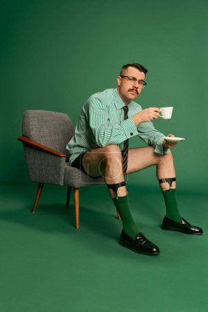 Téléchargez les photos : Morning routine before work. Handsome man, businessman in shirt without pants, sitting on chair, drinking coffee over green background. Concept of emotions, business, occupation, facial expression - en image libre de droit