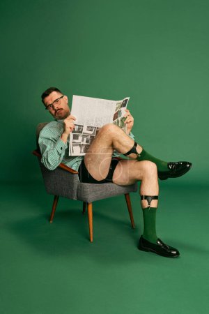 Téléchargez les photos : Portrait of handsome man, in shirt without pants sitting on chair and reading morning press, newspaper posing over green studio background. Concept of emotions, business, occupation, facial expression - en image libre de droit