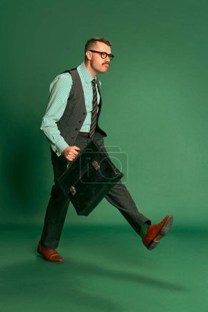 Foto de Portrait of handsome man, businessman in classical suit with briefcase walking to work over green studio background. Concept of emotions, business, occupation, facial expression, fashion - Imagen libre de derechos