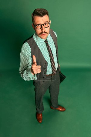 Foto de Top view. Handsome man, businessman in classical suit with briefcase and glasses showing gesture of approvement over green background. Concept of emotions, business, occupation, facial expression - Imagen libre de derechos