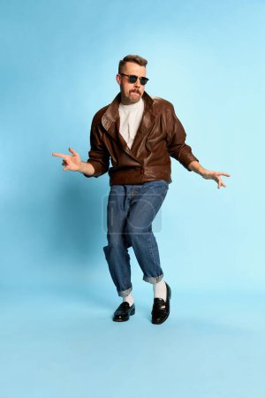 Foto de Joy. Portrait of brutal, handsome man in jeans, leather jacket and sunglasses posing, dancing over blue studio background. Concept of emotions, facial expression, mens fashion, lifestyle. Ad - Imagen libre de derechos