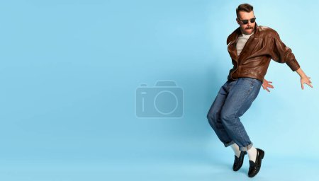 Foto de Portrait of brutal, handsome man in jeans, leather jacket and sunglasses posing, dancing on blue studio background. Banner, flyer. Concept of emotions, facial expression, mens fashion, lifestyle. Ad - Imagen libre de derechos