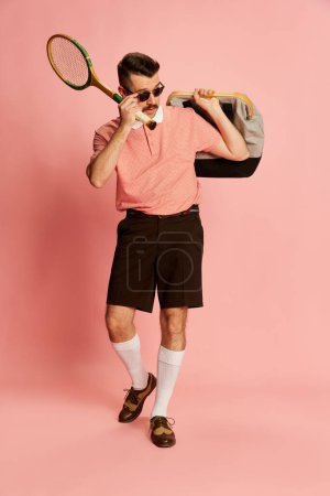 Foto de Portraits of handsome charismatic man in stylish clothes walking with sport bag and tennis racket over pink studio background. Concept of sport, emotions, retro style, lifestyle, fashion. Ad - Imagen libre de derechos