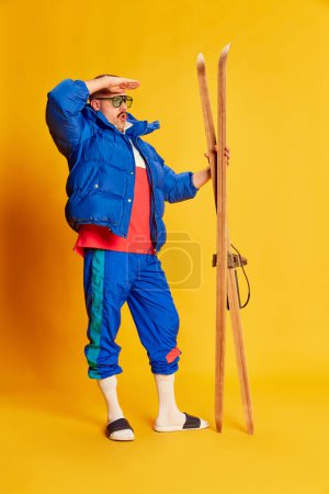 Téléchargez les photos : Active holidays, vacation. Portrait of handsome man in blue winter jacket posing with skis over bright yellow background. Concept of leisure time, winter hobby, emotions, sport, facial expression. Ad - en image libre de droit