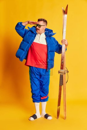 Photo for Portrait of handsome man in blue winter jacket and slippers posing with skis over bright yellow background. Concept of leisure time, winter hobby, emotions, sport, facial expression, fun. Ad - Royalty Free Image