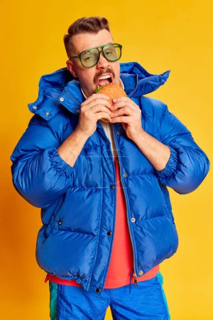 Foto de Delicious. Portrait of handsome man in blue winter jacket posing with burger over bright yellow background. Concept of leisure time, winter holiday, emotions, sport, facial expression, fun. Ad - Imagen libre de derechos