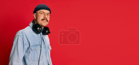 Foto de Portrait of man in stylish modern clothes, glasses, hat and headphones posing over red background. Banner, flyer. Concept of modern fashion, lifestyle, music culture, emotions, facial expression. Ad - Imagen libre de derechos