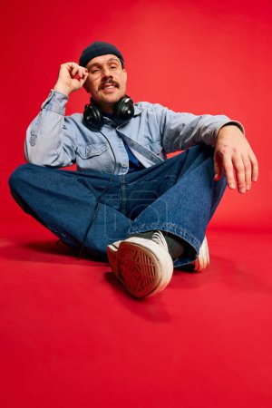 Téléchargez les photos : Portrait of man in stylish modern clothes, oversized jeans, shirt and hat posing over red background. Concept of modern fashion, lifestyle, music culture, emotions, facial expression. Ad - en image libre de droit