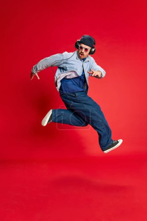 Foto de Party. Portrait of man in stylish clothes, oversized jeans, shirt and hat posing, dancing over red background. Concept of modern fashion, lifestyle, music culture, emotions, facial expression. Ad - Imagen libre de derechos