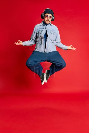 Téléchargez les photos : Relaxation. Portrait of man in stylish clothes, oversized jeans, shirt and hat posing, dancing over red background. Concept of modern fashion, lifestyle, music culture, emotions, facial expression. Ad - en image libre de droit