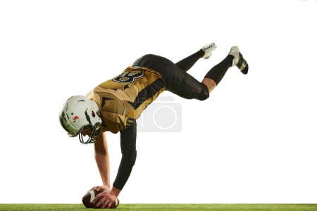Foto de Falling down with ball. Man, american football player in motion, training over white studio background with green grass flooring. Concept of sport, movement, achievements, competition, hobby - Imagen libre de derechos
