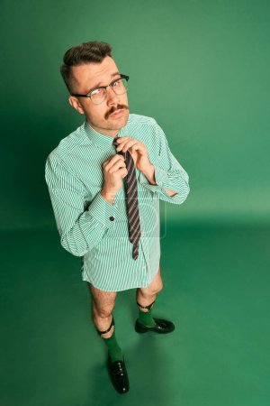 Photo for Top view. Portrait of handsome man, businessman in shirt without pants, tying tie, posing over green studio background. Concept of emotions, business, occupation, facial expression, fashion - Royalty Free Image