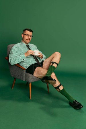 Téléchargez les photos : Portrait of handsome, brutal man, businessman in shirt without pants sitting on chair and drinking morning coffee over green background. Concept of emotions, business, occupation, facial expression - en image libre de droit
