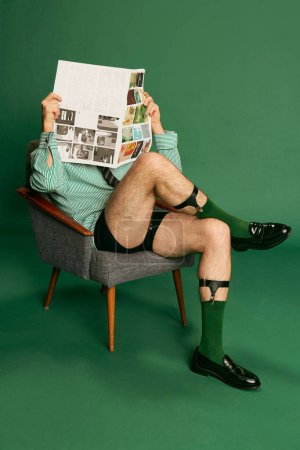 Téléchargez les photos : Portrait of man, businessman in shirt without pants sitting on chair and reading newspaper, posing over green studio background. Concept of emotions, business, occupation, facial expression, fashion - en image libre de droit