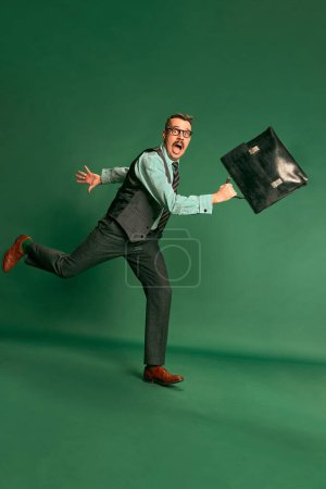 Téléchargez les photos : Portrait of handsome man, businessman in classical suit with briefcase actively running to work over green studio background. Concept of emotions, business, occupation, facial expression, fashion - en image libre de droit
