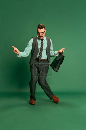 Foto de Cheerful. Portrait of handsome man, businessman in classical suit with briefcase dancing over green studio background. Concept of emotions, business, occupation, facial expression, fashion - Imagen libre de derechos