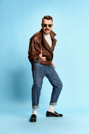Photo for Portrait of brutal, handsome man in jeans, leather jacket and sunglasses posing, dancing over blue studio background. Confidence. Concept of emotions, facial expression, mens fashion, lifestyle. Ad - Royalty Free Image