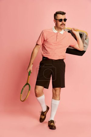 Foto de Sport club member. Portraits of handsome charismatic man in stylish clothes posing with tennis racket over pink studio background. Concept of sport, emotions, retro style, lifestyle, fashion. Ad - Imagen libre de derechos