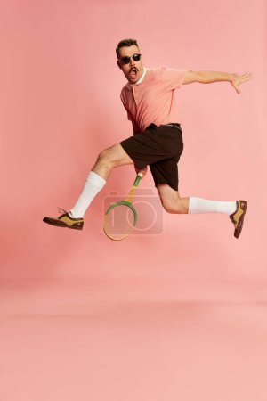 Foto de Portraits of handsome charismatic man in stylish clothes posing with tennis racket in a jump over pink studio background. Concept of sport, emotions, retro style, lifestyle, fashion. Ad - Imagen libre de derechos