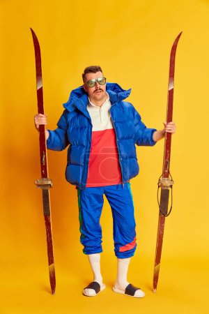 Foto de Portrait of handsome man in blue winter jacket posing with skis over bright yellow background. Holiday activity. Concept of leisure time, winter hobby, emotions, sport, facial expression, fun. Ad - Imagen libre de derechos