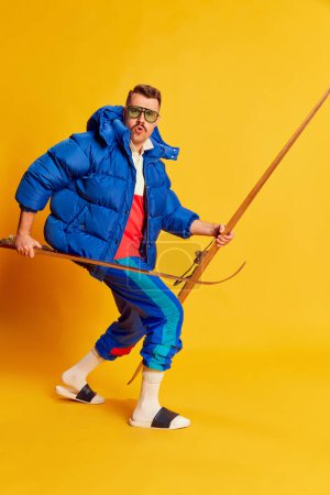 Foto de Portrait of handsome man in blue winter jacket posing with skis over bright yellow background. Sportsman. Concept of leisure time, winter hobby, emotions, sport, facial expression, fun. Ad - Imagen libre de derechos