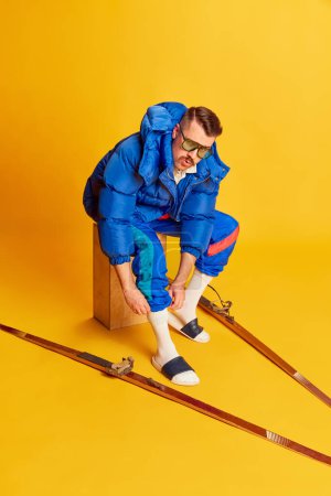 Téléchargez les photos : Emotive face. Portrait of handsome man in blue winter jacket posing with skis over bright yellow background. Concept of leisure time, winter hobby, emotions, sport, facial expression, fun. Ad - en image libre de droit
