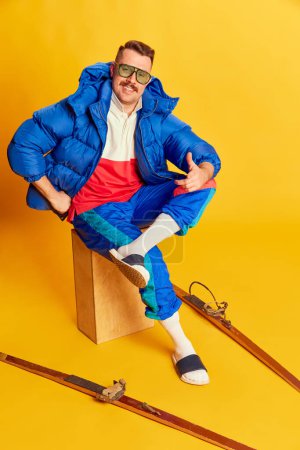 Téléchargez les photos : Positive mood. Portrait of handsome man in blue winter jacket posing with skis over bright yellow background. Concept of leisure time, winter hobby, emotions, sport, facial expression, fun. Ad - en image libre de droit