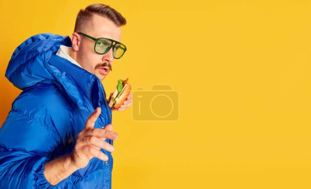 Foto de Portrait of handsome man in blue winter jacket posing with burger over bright yellow background. Banner, flyer. Concept of leisure time, winter holiday, emotions, sport, facial expression, fun. Ad - Imagen libre de derechos