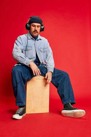 Foto de Music in headphones. Man in stylish modern clothes, oversized jeans, shirt and hat posing over red background. Concept of modern fashion, lifestyle, music culture, emotions, facial expression. Ad - Imagen libre de derechos