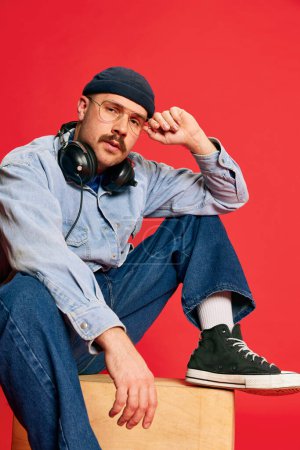 Photo for Portrait of man in stylish clothes, oversized jeans, shirt, hat and headphones posing over red background. Concept of modern fashion, lifestyle, music culture, emotions, facial expression. Ad - Royalty Free Image