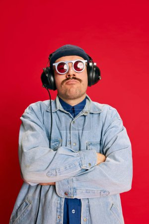 Téléchargez les photos : Portrait of man in jeans shirt, sunglasses, listening to music in headphones, posing over red background. Concept of modern fashion, lifestyle, music culture, emotions, facial expression. Ad - en image libre de droit