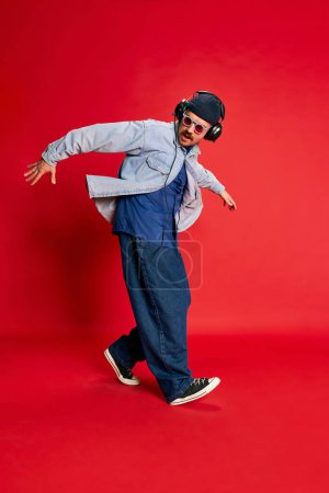 Foto de Hip hop. Portrait of man in stylish clothes, oversized jeans, shirt and hat posing, dancing over red background. Concept of modern fashion, lifestyle, music culture, emotions, facial expression. Ad - Imagen libre de derechos