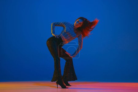 Téléchargez les photos : Dynamics. Portrait of young girl dancing high heel dance in stylish clothes over blue background in neon light. Concept of dance lifestyle, modern style, contemporary, youth culture, self-expression - en image libre de droit