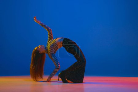 Téléchargez les photos : Flexibility. Portrait of young girl dancing high heel dance in stylish clothes on blue background in neon light. Concept of dance lifestyle, modern style, contemporary, youth culture, self-expression - en image libre de droit