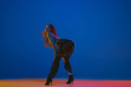 Téléchargez les photos : Femininity. Portrait of young girl dancing high heel dance in stylish clothes over blue background in neon light. Concept of dance lifestyle, modern style, contemporary, youth culture, self-expression - en image libre de droit